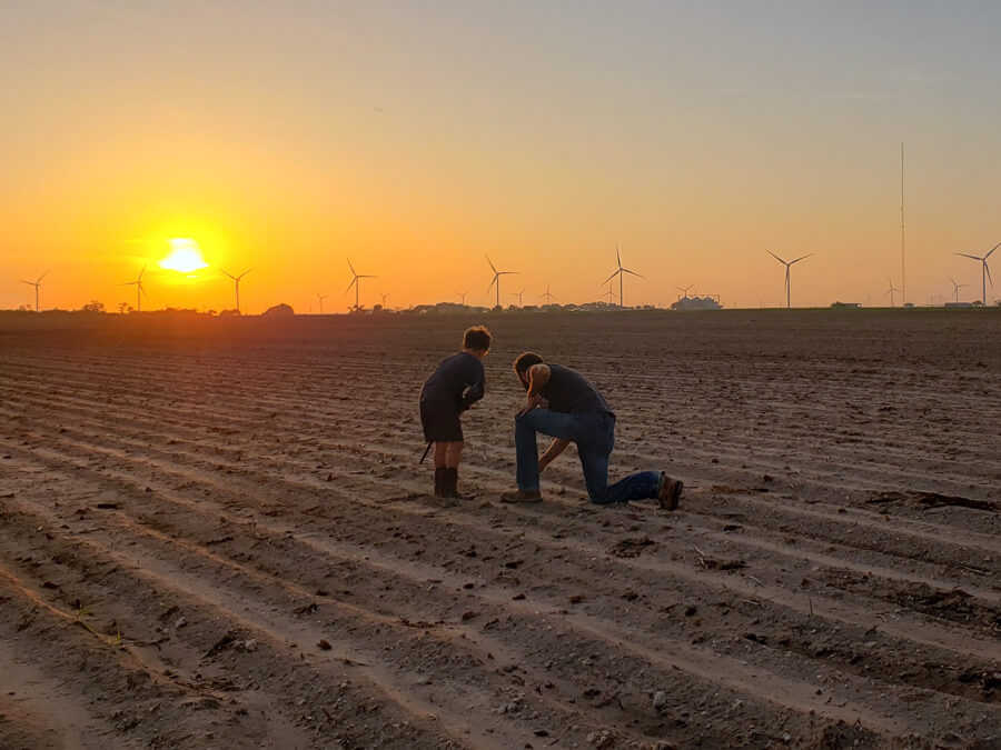 Farmer and son inspecting the soil at sunrise with wind turbines in the background