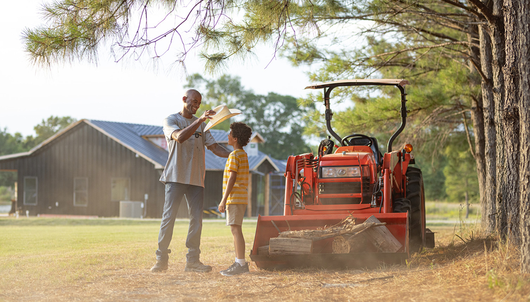 Father placing cowboy hat on young son by a tractor